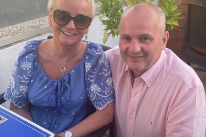 Pauline and Declan sitting in a restaurant with his arm around her smiling