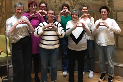 A group of 8 women all standing making a heart symbol with their hands