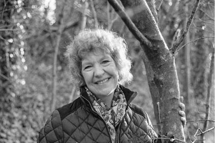 A black and white photo of Gill smiling, she has short curly hair and is wearing a dark coloured quilted jacket