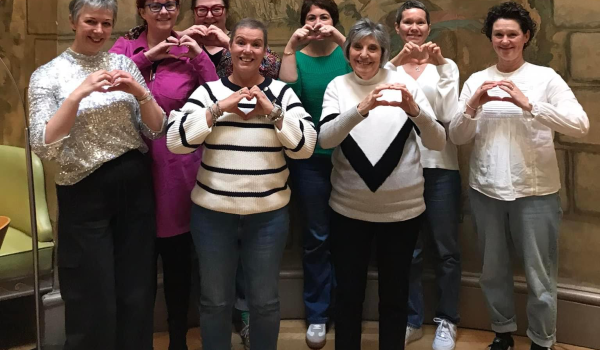 A group of 8 women all standing making a heart symbol with their hands