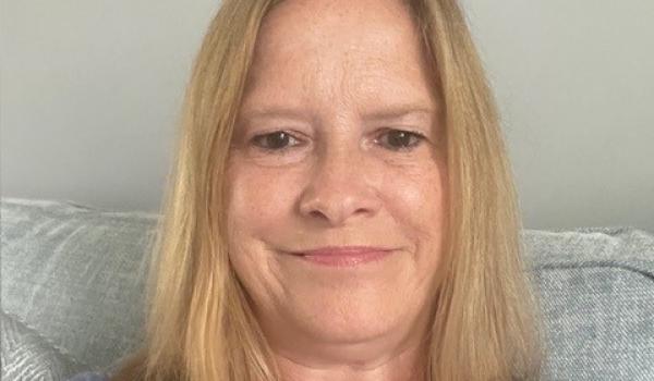 A selfie that Judy is taking sitting on her sofa wearing a blue patterned top with her blonde hair worn down and she's smiling