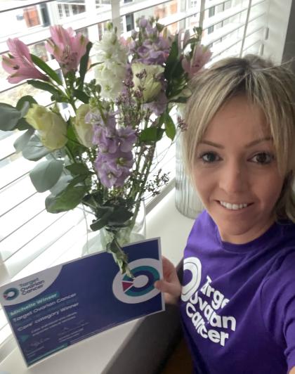 A photo of Michelle Winser with her Nurse Hero Award certificate and flowers wearing a Target Ovarian Cancer tshirt