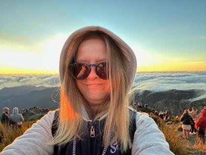 A photo of Judy standing on top of the mountain with the sunrise behind her. She's smiling, with her hood up and wearing sunglasses