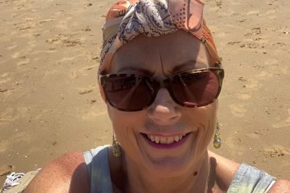 Jeanette taking a selfie on the beach, smiling wearing sunglasses and a headscarf