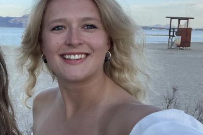 Lotte standing on the beach taking a selfie with wavy blonde hair and wearing a white top