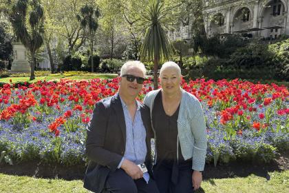 Wendy and her husband both standing together and smiling for a photo outside a colourful flower bed