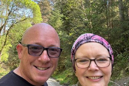 A selfie of Lesley and her partner out for a walk in the sun smiling. They are both wearing glasses and Lesley is wearing a scarf on her head