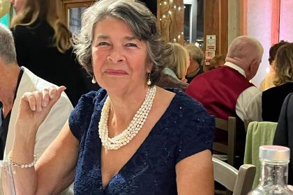 A photo of Georgina sitting at a table smiling. She is wearing a dark coloured dress and a cream necklace