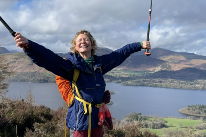 Janet standing on top of a hill with a huge smile, she's holding both of her hands out and up in the air in celebration holding two walking sticks as well