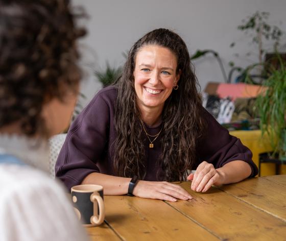 A photo of Lindsay sitting at a table with her hands resting on it smiling. She has long brown curly hair and is wearing a brown top