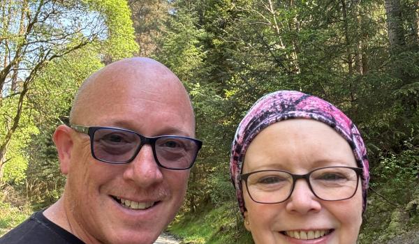 A selfie of Lesley and her partner out for a walk in the sun smiling. They are both wearing glasses and Lesley is wearing a scarf on her head