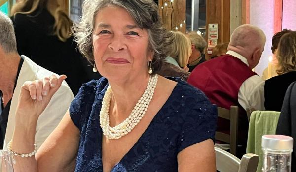 A photo of Georgina sitting at a table smiling. She is wearing a dark coloured dress and a cream necklace