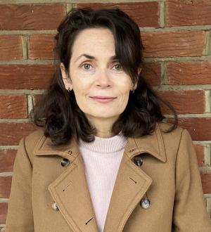 Elaine standing in front of a brick wall wearing a camel colour trench coat and peach top