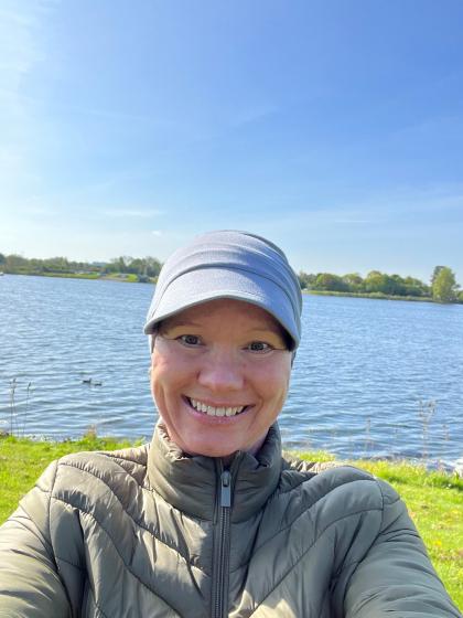 Sarah taking a selfie wearing a cap and grey jacket, she's outside in front of a lake with blue skies behind her