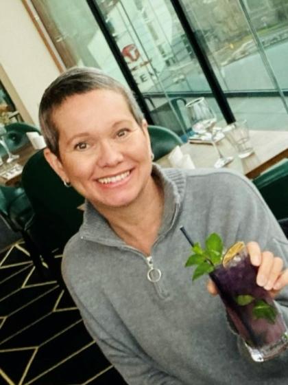 Sarah with short hair wearing a grey jumper with a drink in her hand smiling