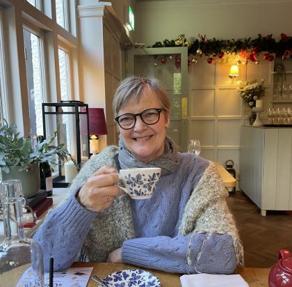 Nicola sitting in a cafe drinking a cup of tea wearing black glasses and a cosy jumper smiling for a photo