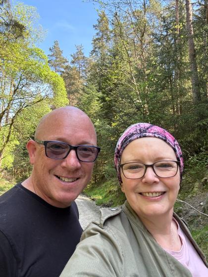 A selfie of Lesley and her partner out for a walk in the sun smiling. They are both wearing glasses and Lesley is wearing a scarf on her head