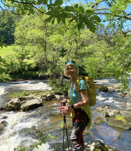 Janet standing next to a stream surrounded by trees wearing walking clothes and carry two walking sticks. She's smiling for the camera