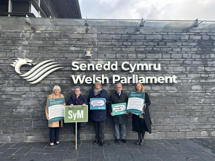 Campaigners with placards in Welsh and English about ovarian cancer, standing in front of wall with writing on it stating that it's the Senedd